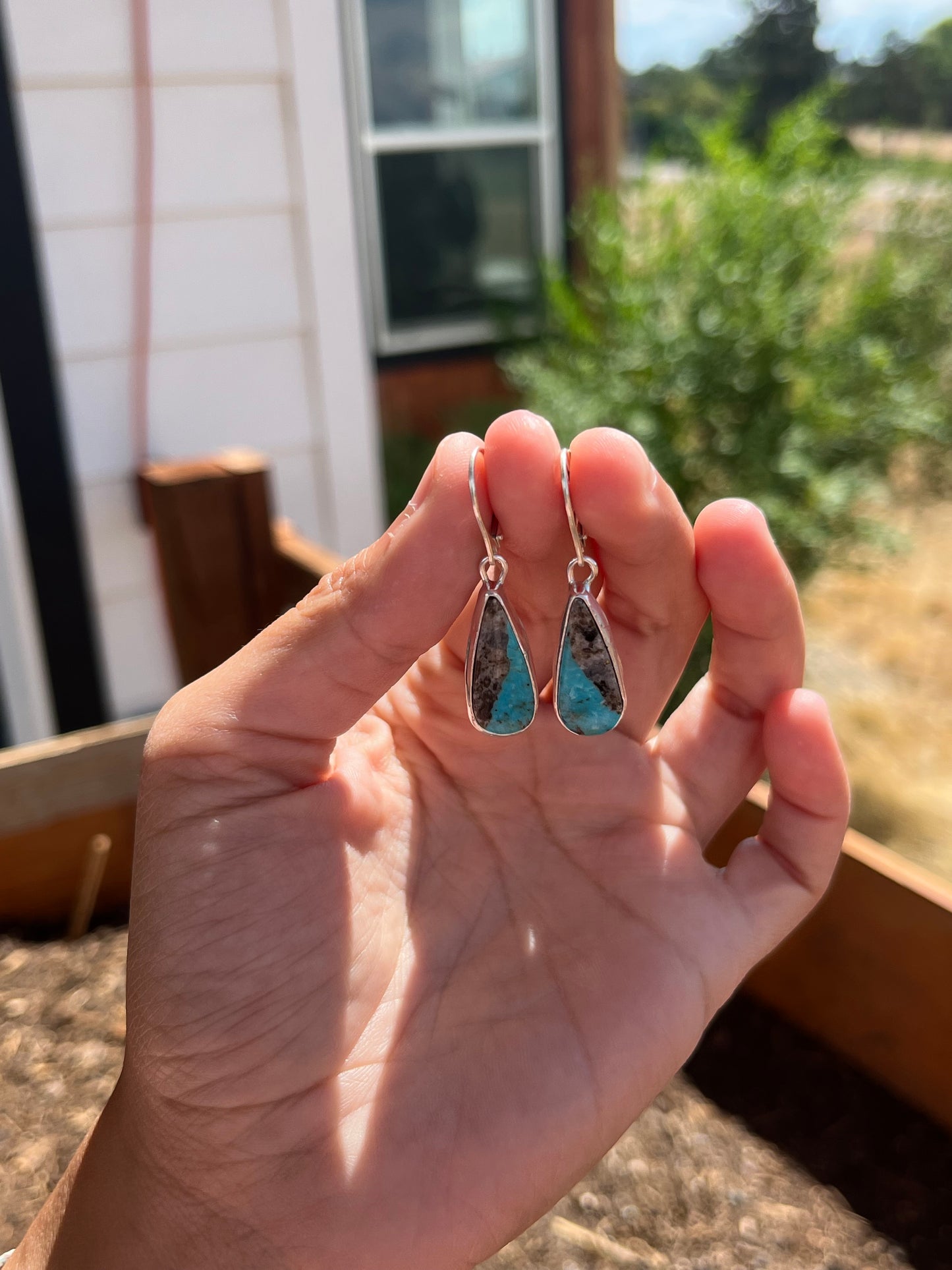 Hand holding two turquoise teardrop-shaped earrings with a blurred outdoor background