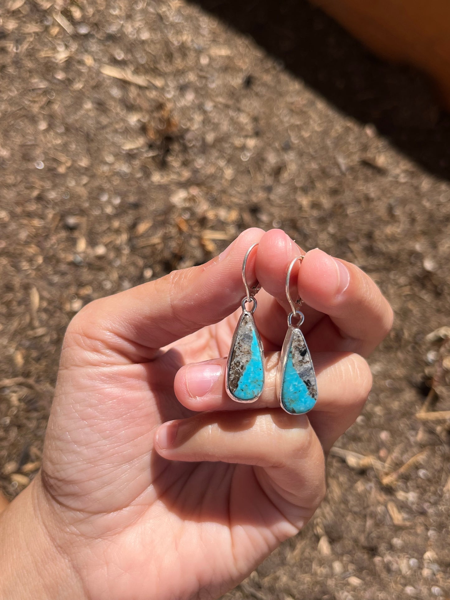 Hand holding two turquoise teardrop-shaped earrings on a textured surface.